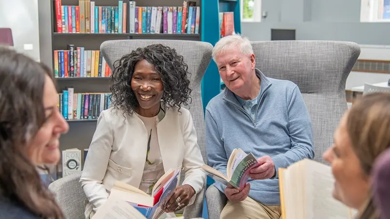 Adults of various ages sitting together in a library, smiling and holding books during what appears to be a reading group discussion.