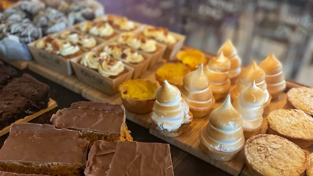 Assorted cakes and pastries displayed on wooden boards, including chocolate slices, cream-topped meringues, and biscuits.