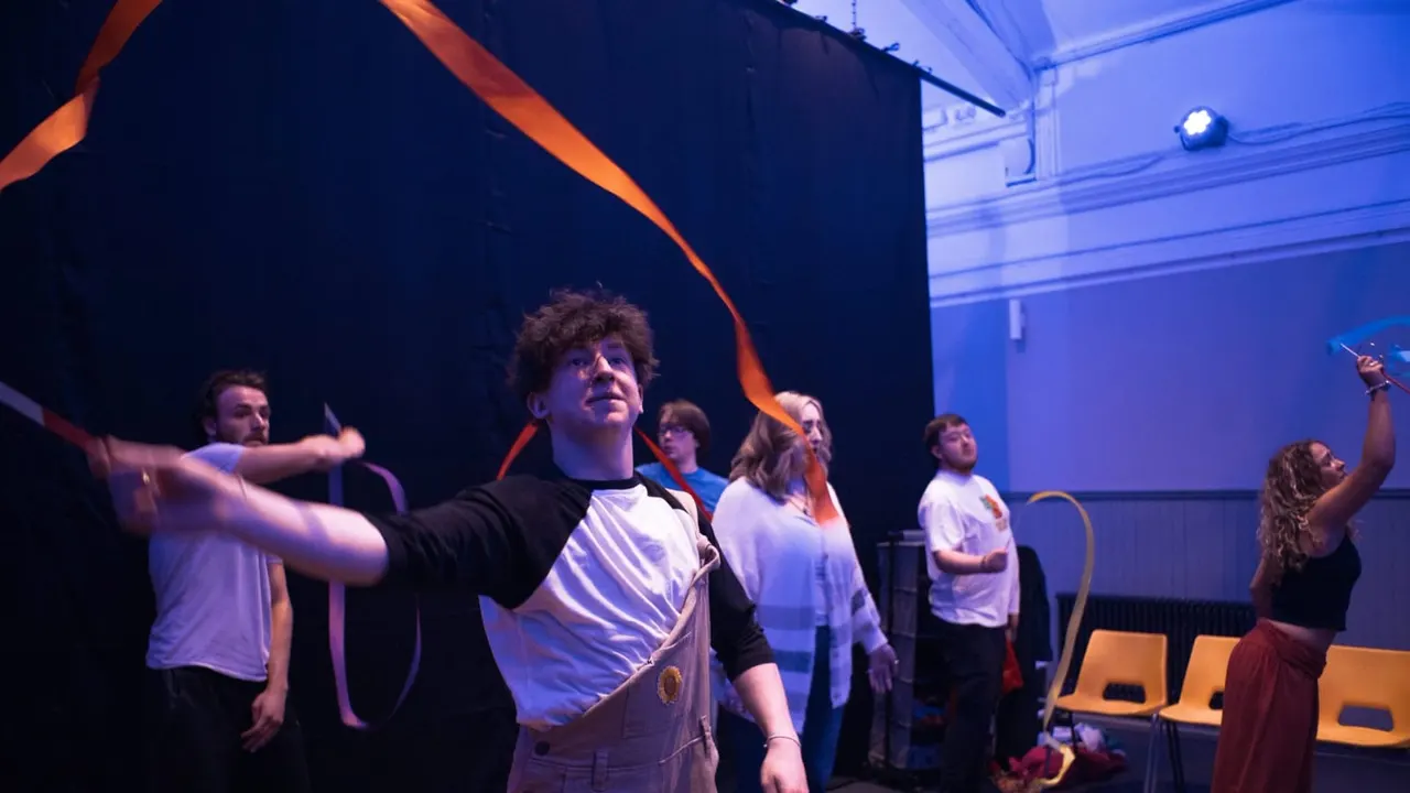 Group of young people performing on stage with ribbon props under blue and orange lighting.