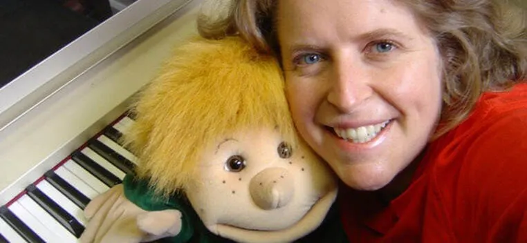 Woman smiling with a puppet character with yellow fur at a piano keyboard.
