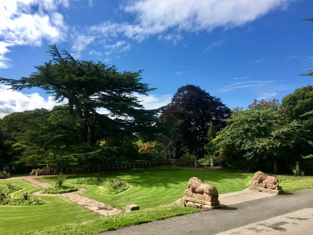 Manicured gardens with stone lion sculptures, mature trees including a windswept cedar, and a stone balustrade under blue sky.