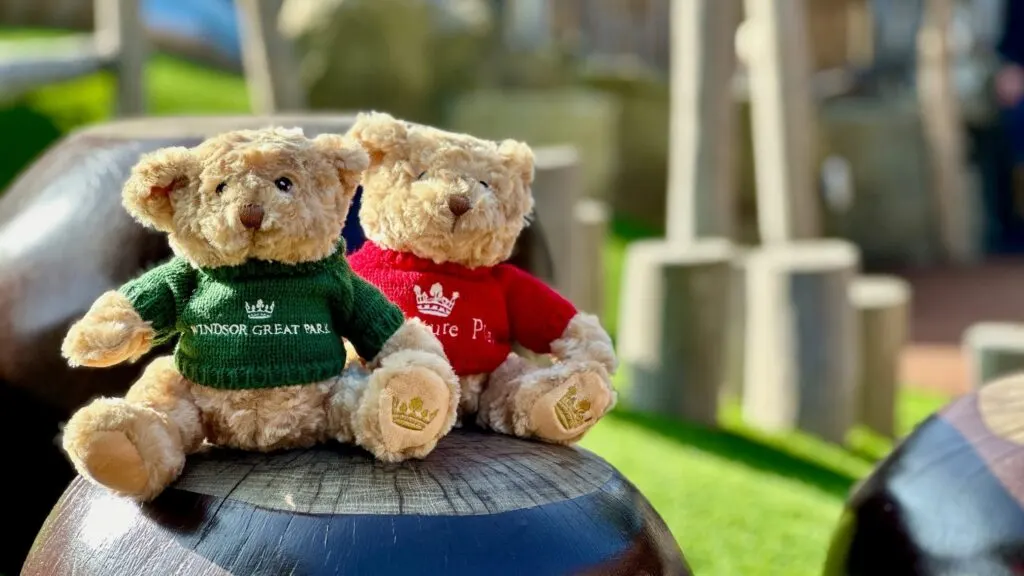 Two teddy bears wearing branded sweaters sit on a wooden post in a garden setting with green lawn and fencing visible behind.