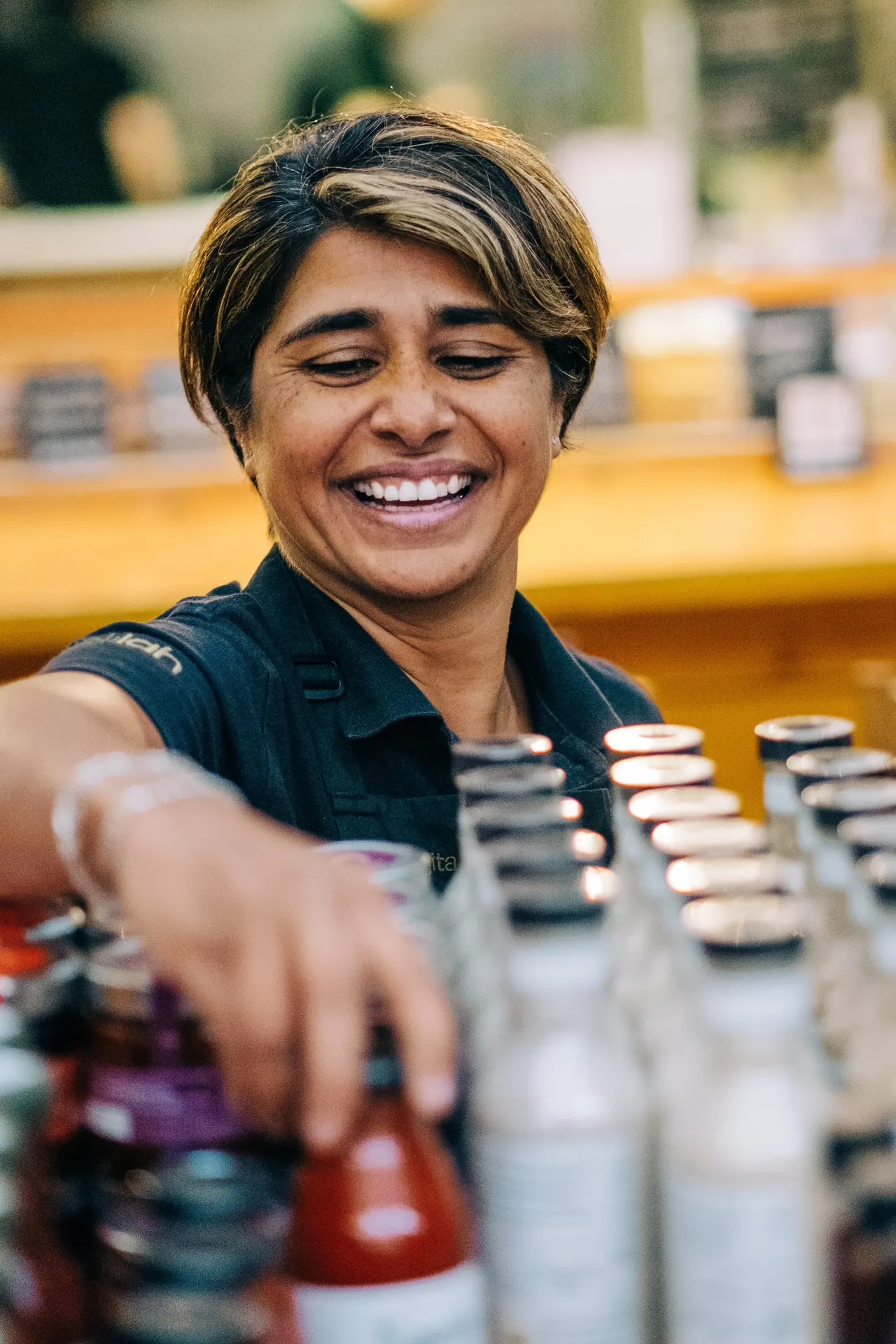 A smiling staff member in a dark polo shirt reaching towards bottles behind a counter at a food venue.
