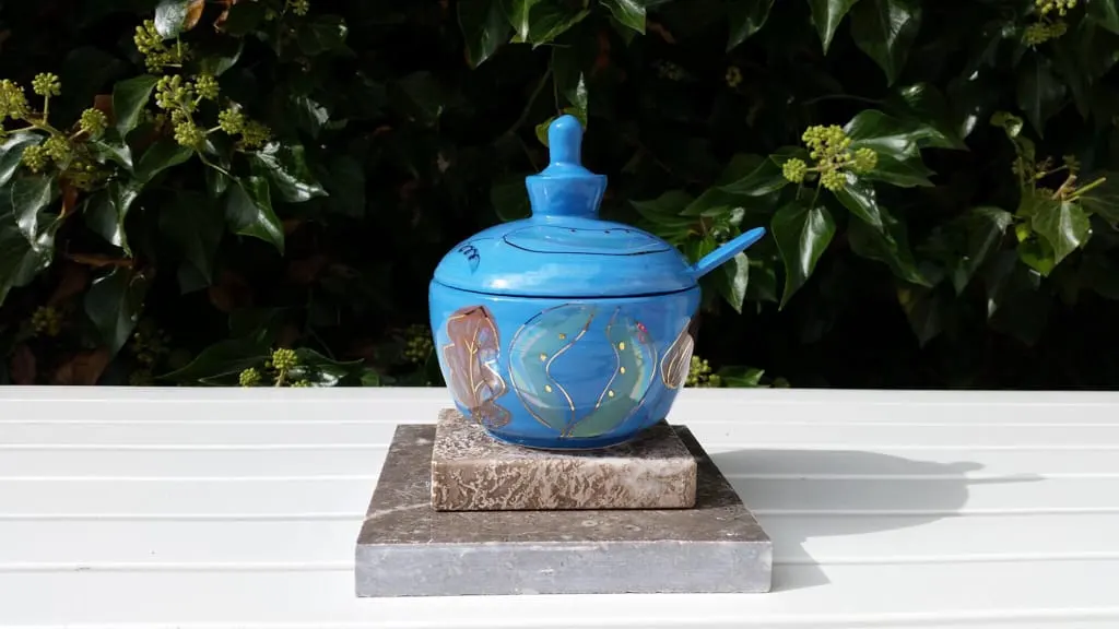 Bright blue ceramic teapot with decorative hand-painted details displayed on a stone base against leafy garden foliage.