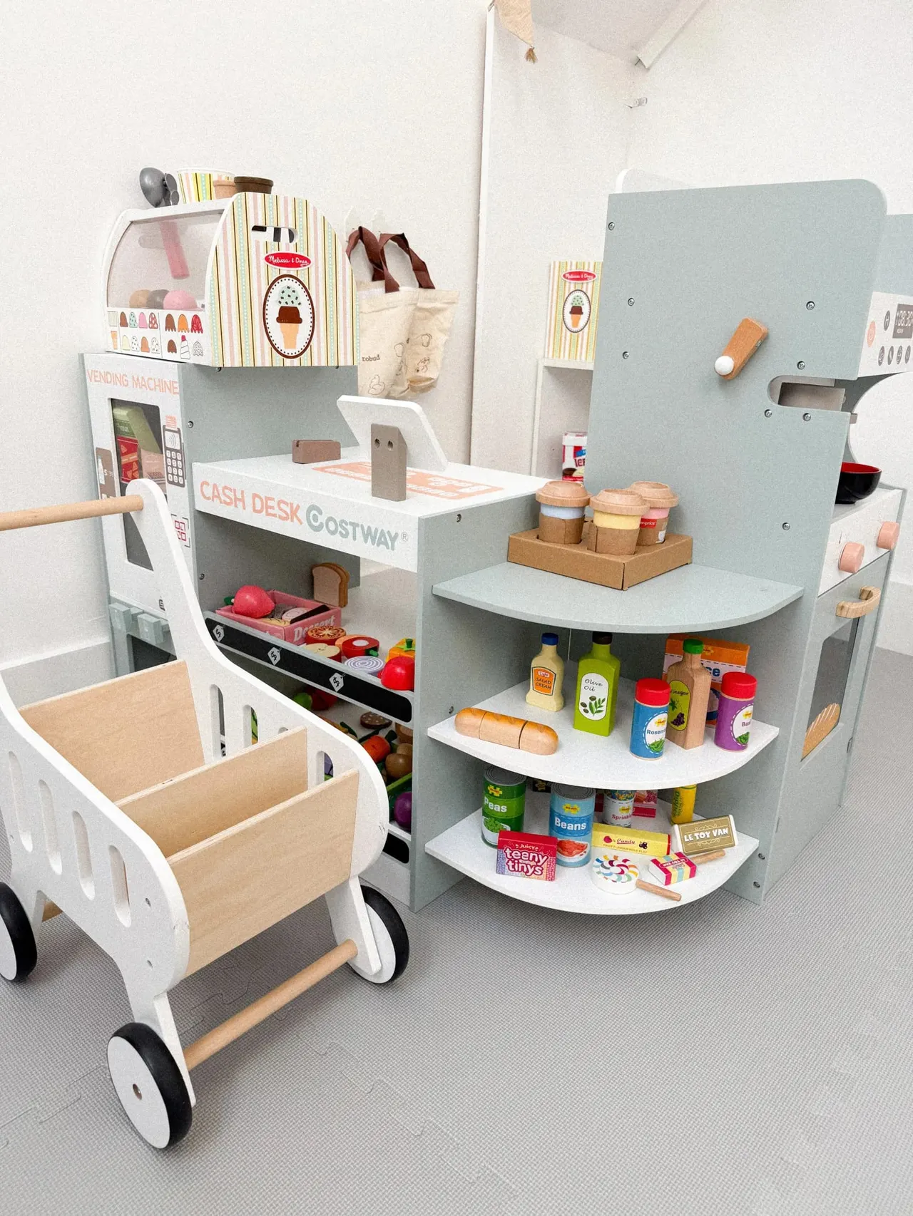 Wooden toy shop and café playset with a shopping trolley, cash desk, shelving stocked with pretend groceries and drinks bottles, and ice-cream counter.