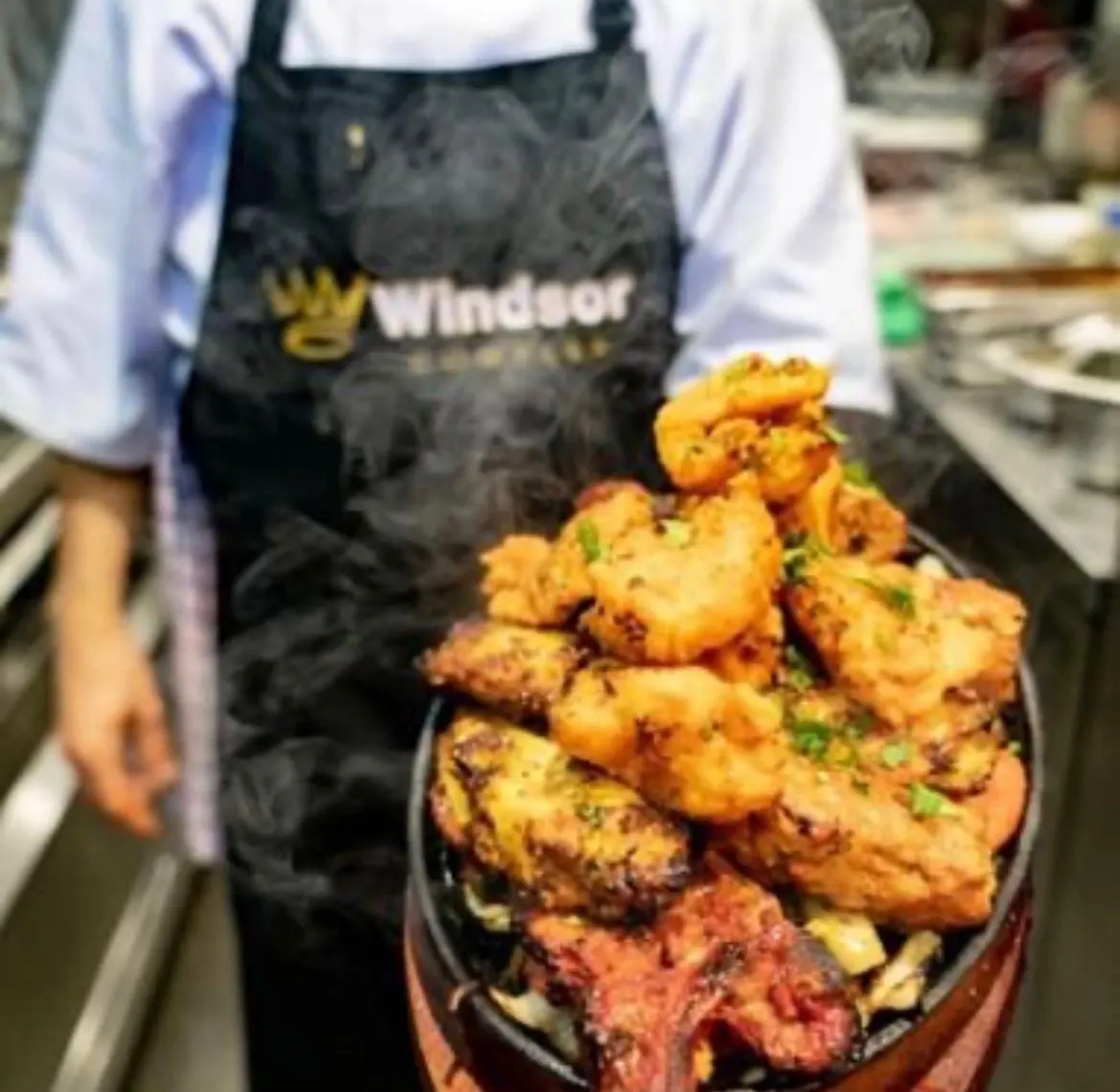 Chef holding a platter of golden-fried prawns and meat garnished with herbs in a kitchen setting.