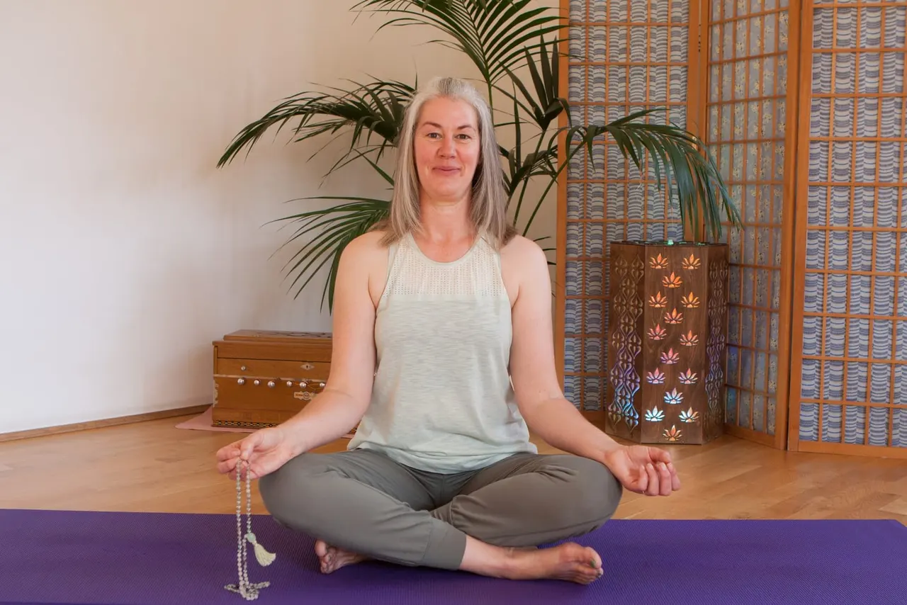 Woman sitting cross-legged on a purple yoga mat in a meditation pose, holding prayer beads, surrounded by plants and wooden décor.