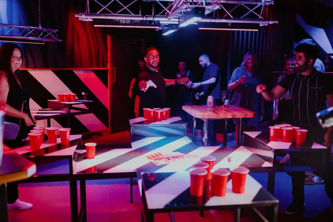Group of people playing beer pong at tables in a brightly lit indoor venue with red and blue neon lighting.