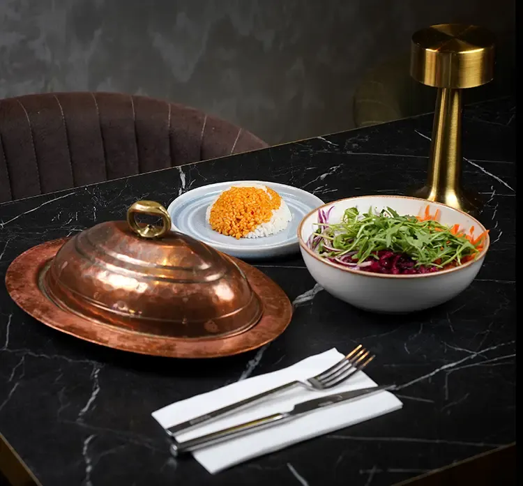 Plated meal with covered copper dome, rice dish, and fresh salad on black marble table with place setting.