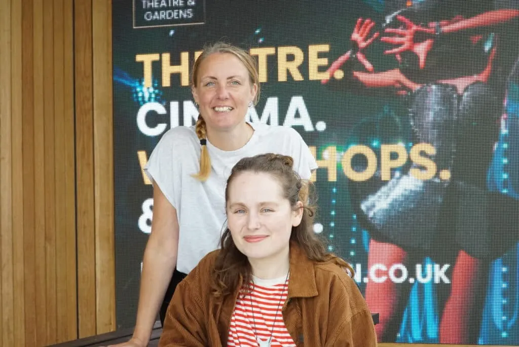 Two women posing together in front of a theatre and cinema promotional screen display.
