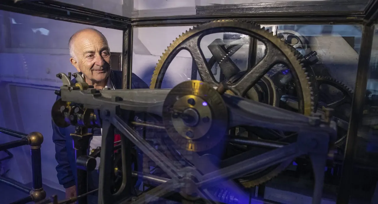 An older man examines a large brass gear and mechanical machinery on display inside a glass case.