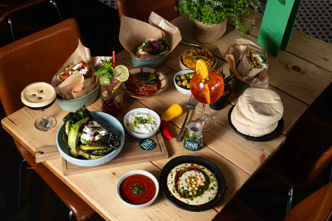 Overhead view of a wooden table laden with Mediterranean mezze dishes, cocktails, and appetisers at a restaurant.