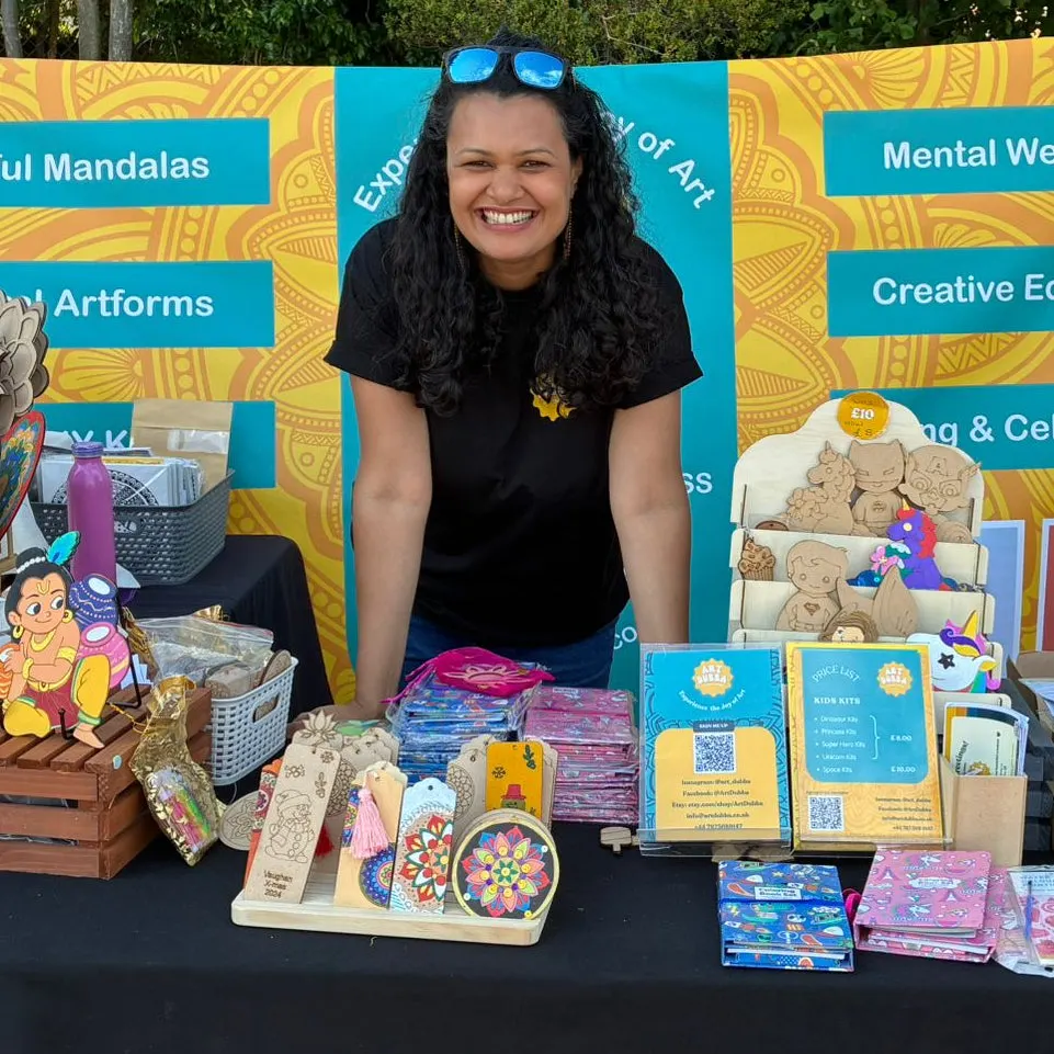 Woman smiling behind a market stall displaying colourful wooden art and craft items including mandalas and decorative boxes.