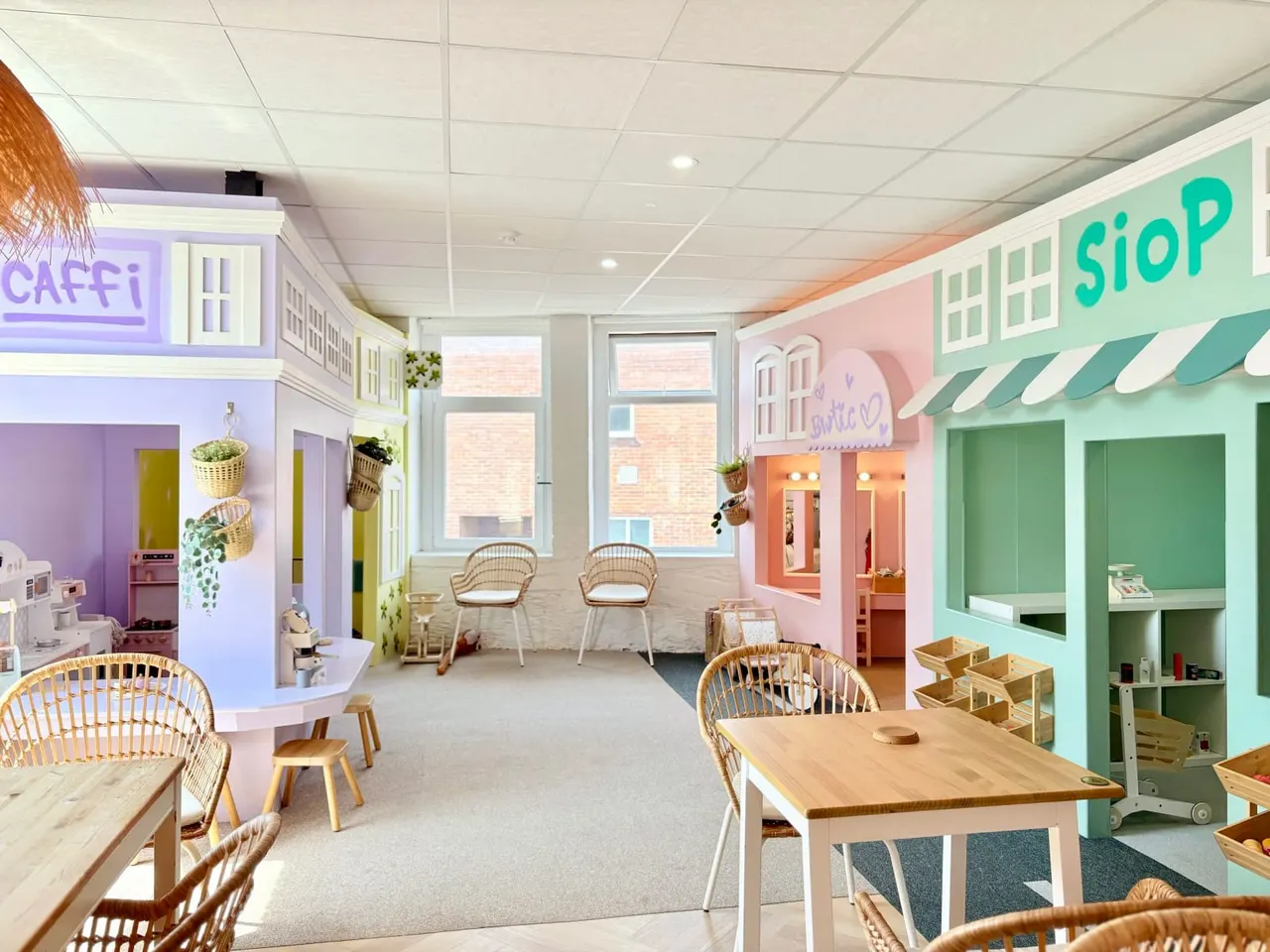 Bright, colourful indoor play space with themed shop and café sets, wooden tables, wicker chairs, and play structures in purple, pink and green.
