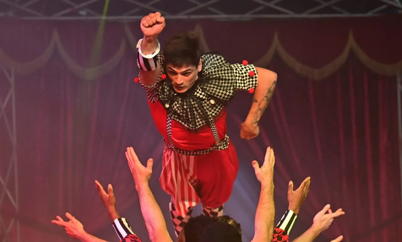 Circus performer in red and black harlequin costume jumping above audience members with raised hands.