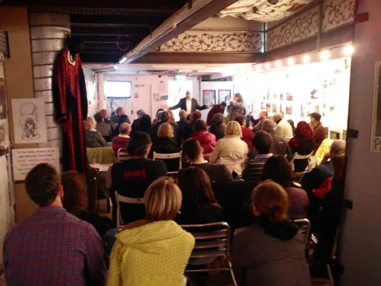 A full audience seated indoors watching a presentation or performance at a small venue with illuminated display panels on the walls.