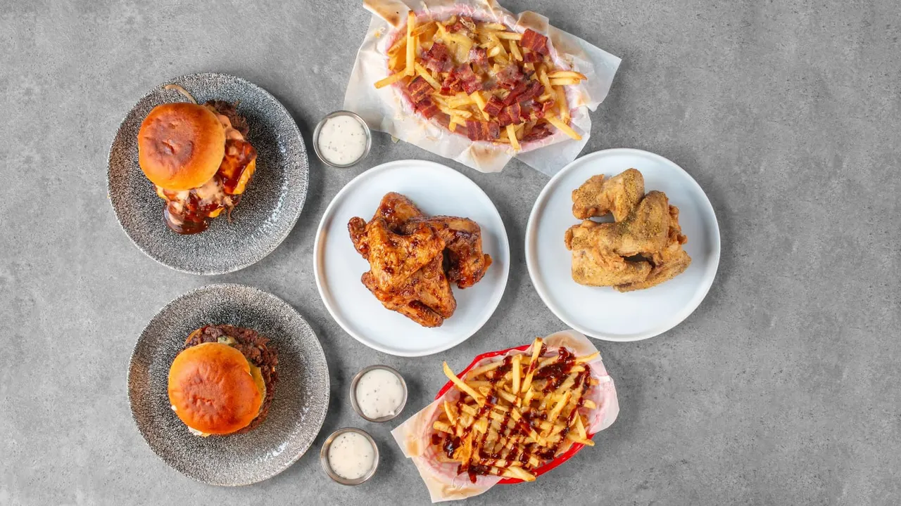 Overhead view of various dishes including burgers, loaded chips with bacon, and seasoned chicken wings on a grey surface.