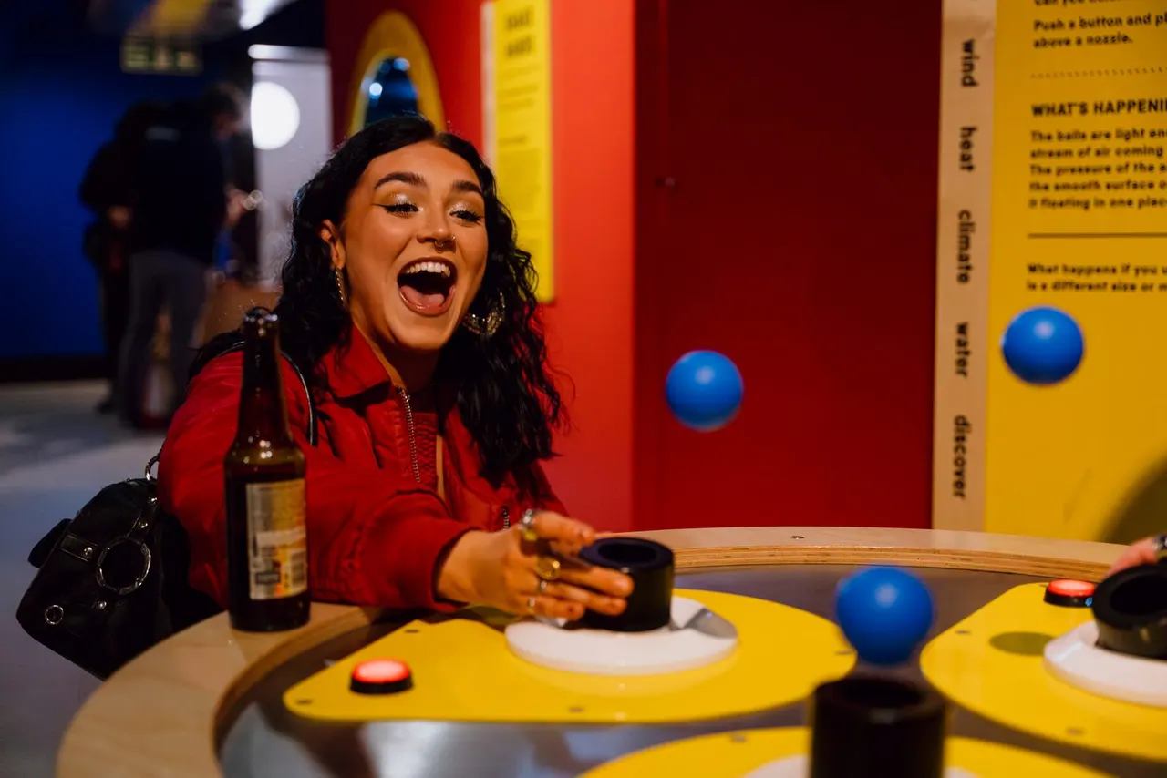 Woman in red jacket laughing whilst interacting with a colourful interactive science exhibit with blue balls and yellow panels.