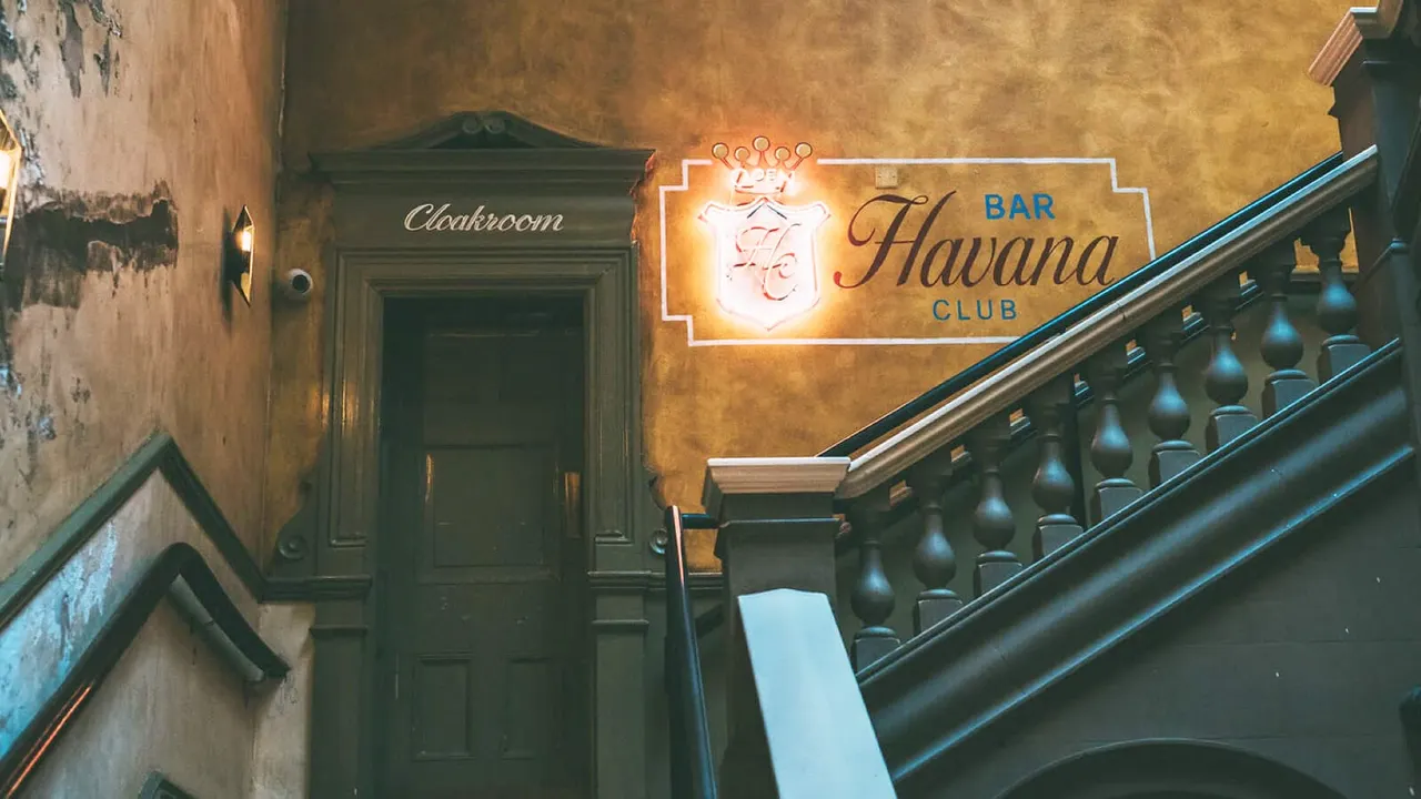 A neon-lit "Havana Club Bar" sign glows above a staircase with ornate black railings in a vintage building with weathered ochre walls.
