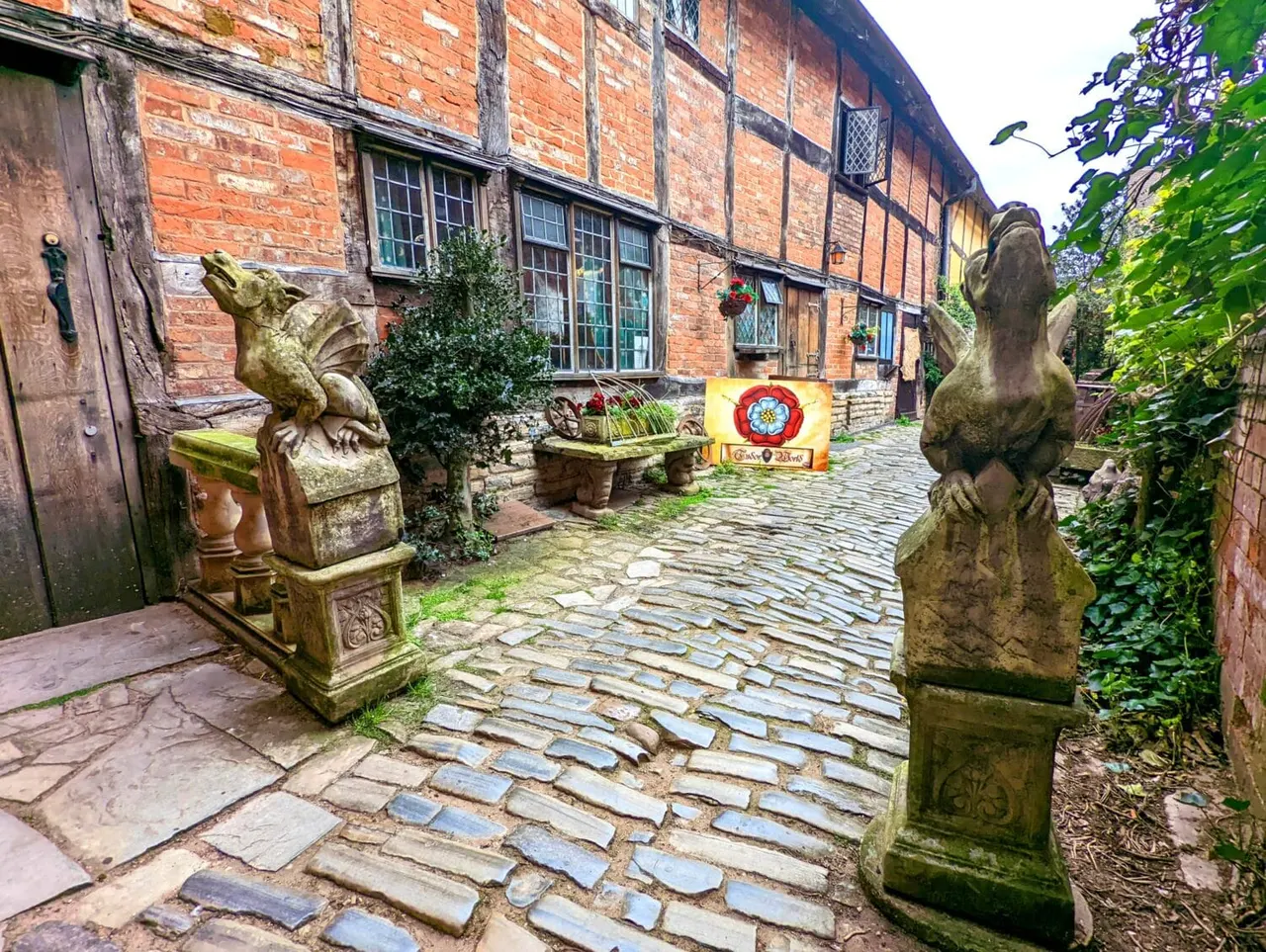 Stone courtyard with Tudor building, stone bird sculptures, climbing ivy, and decorative planters.