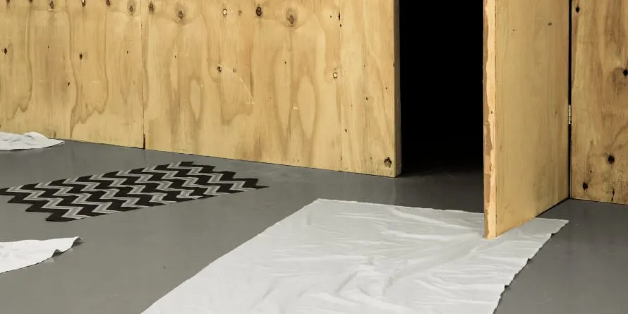 Interior of empty gallery space with plywood walls, concrete floor, and white plastic sheeting.
