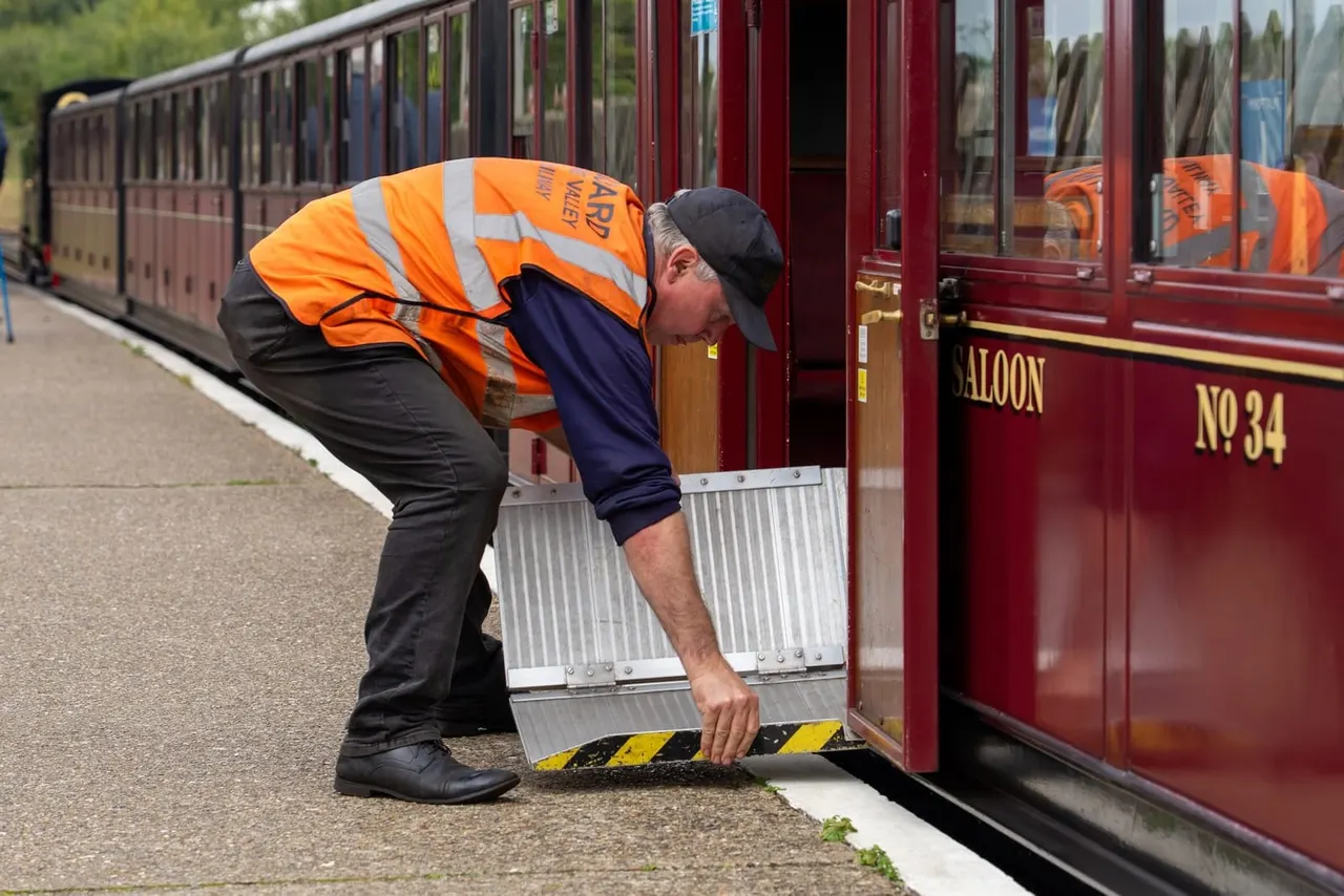 A railway worker in high-visibility clothing deploys a portable ramp at the door of a heritage train carriage for passenger access.
