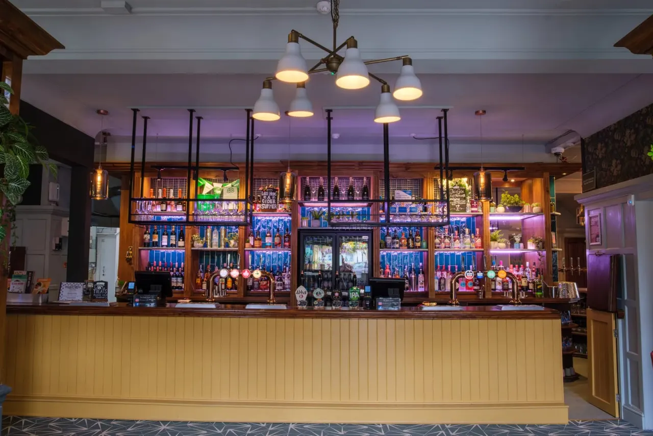 A spacious bar interior with a long cream-coloured counter, extensive shelving displaying spirits and beers, brass beer taps, and purple ambient lighting from above.