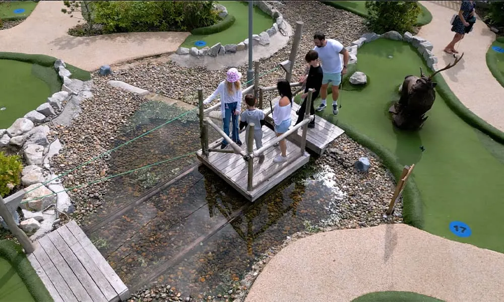 Overhead view of visitors playing mini golf on a course with a pond and wooden platform bridge.