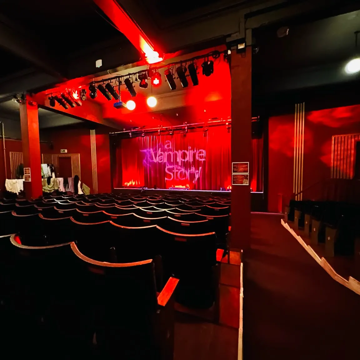 Empty theatre auditorium with red lighting, rows of black seats, and a stage with neon signage.