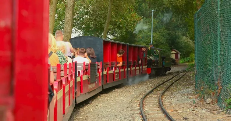 A miniature railway train with red and yellow carriages carrying passengers along a curved track through woodland.