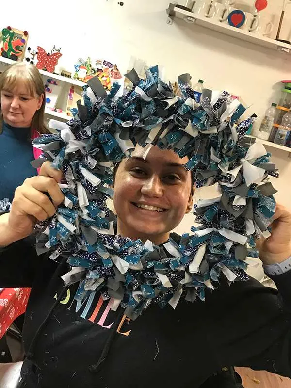 Two children wearing a large decorative wreath made of blue, white and patterned fabric ribbons in a craft room.