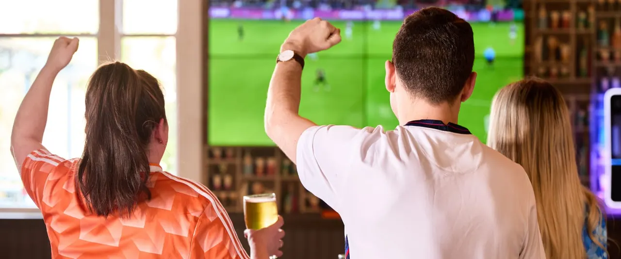 Three people watching a football match on a large screen in a pub, celebrating with raised fists.