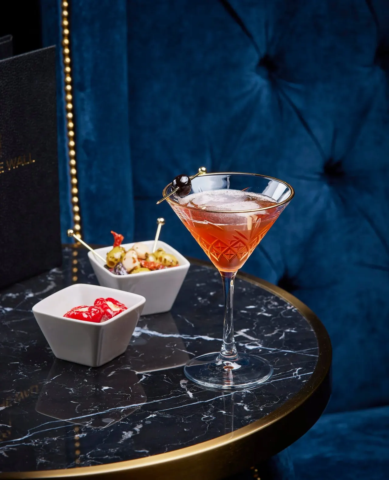 Cocktail with cherry garnish and appetisers on a marble table against a blue velvet backdrop.