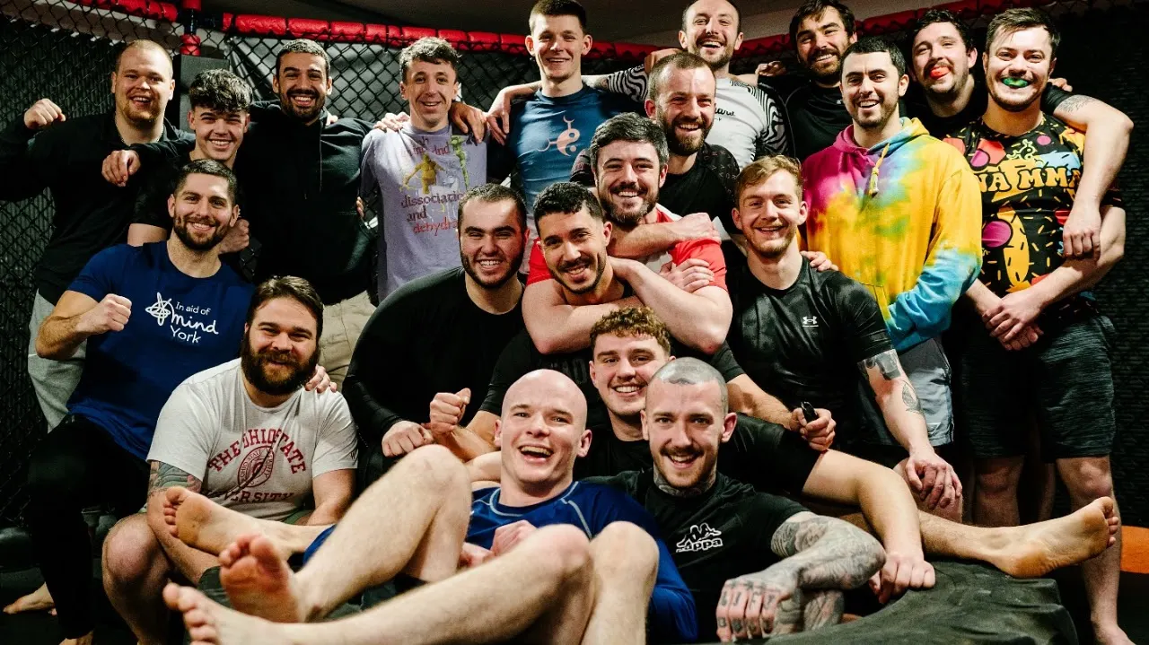 Large group of men posing together inside a boxing or MMA gym with red neon lighting and a chain-link fence backdrop.