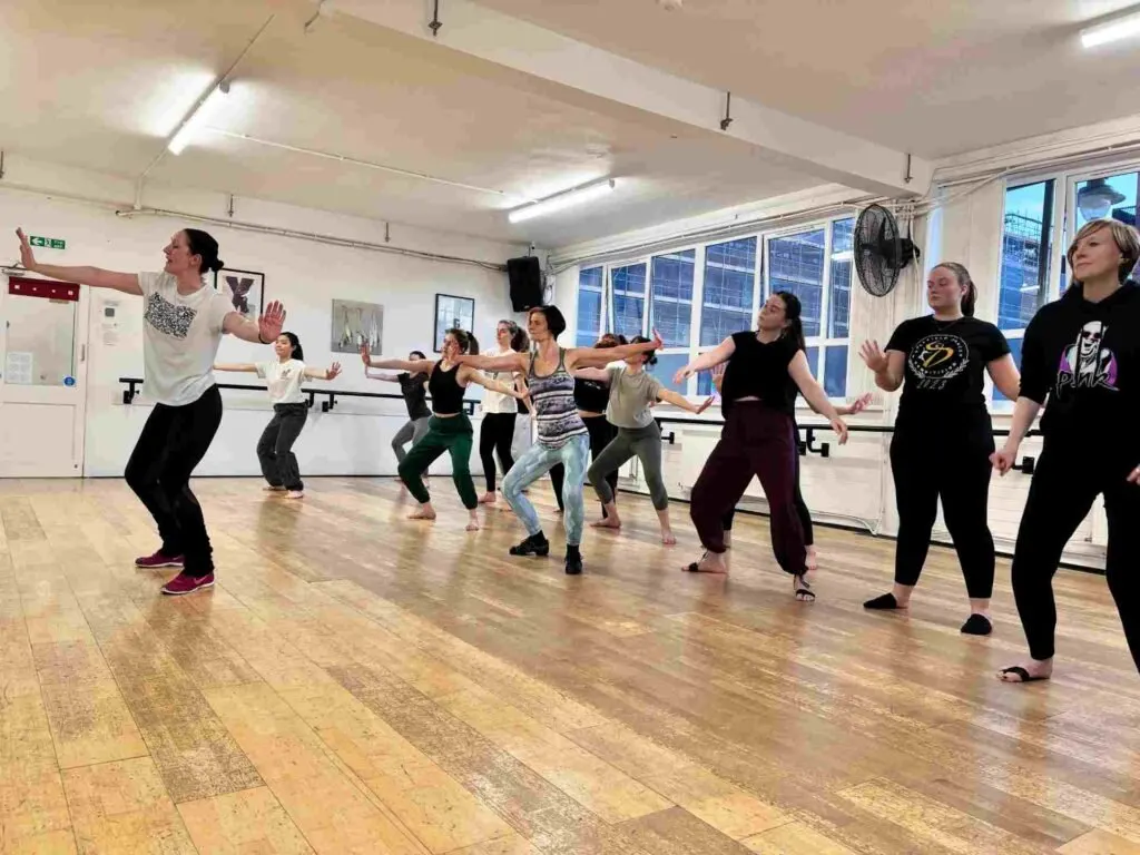 A group of people of mixed ages participating in a dance class in a studio with wooden flooring and mirrored walls.