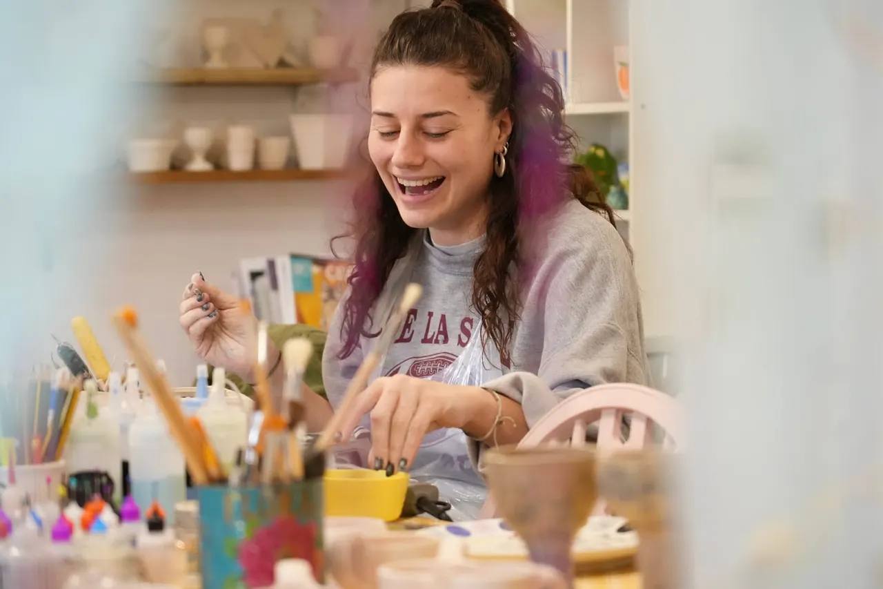 A woman smiling whilst working at a pottery crafting table surrounded by art supplies and materials.