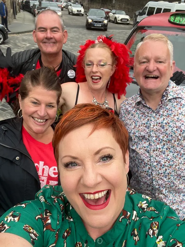 Five people smiling at camera on a street, some wearing red wigs and festive clothing.