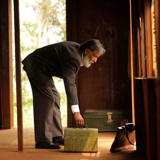 Older man in dark suit bending over a vintage suitcase in a wooden shelter with open doors and trees visible beyond.
