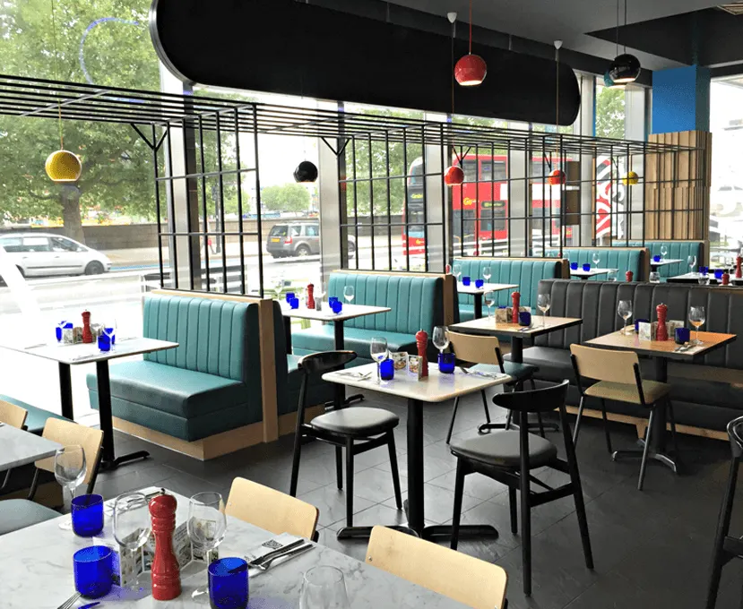 Modern restaurant interior with turquoise booth seating, tables with blue glassware, and large windows overlooking a street.