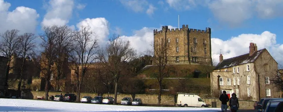 A stone castle keep dominates a hilltop overlooking a snowy foreground with parked cars, bare trees, and adjacent stone buildings on a winter's day.