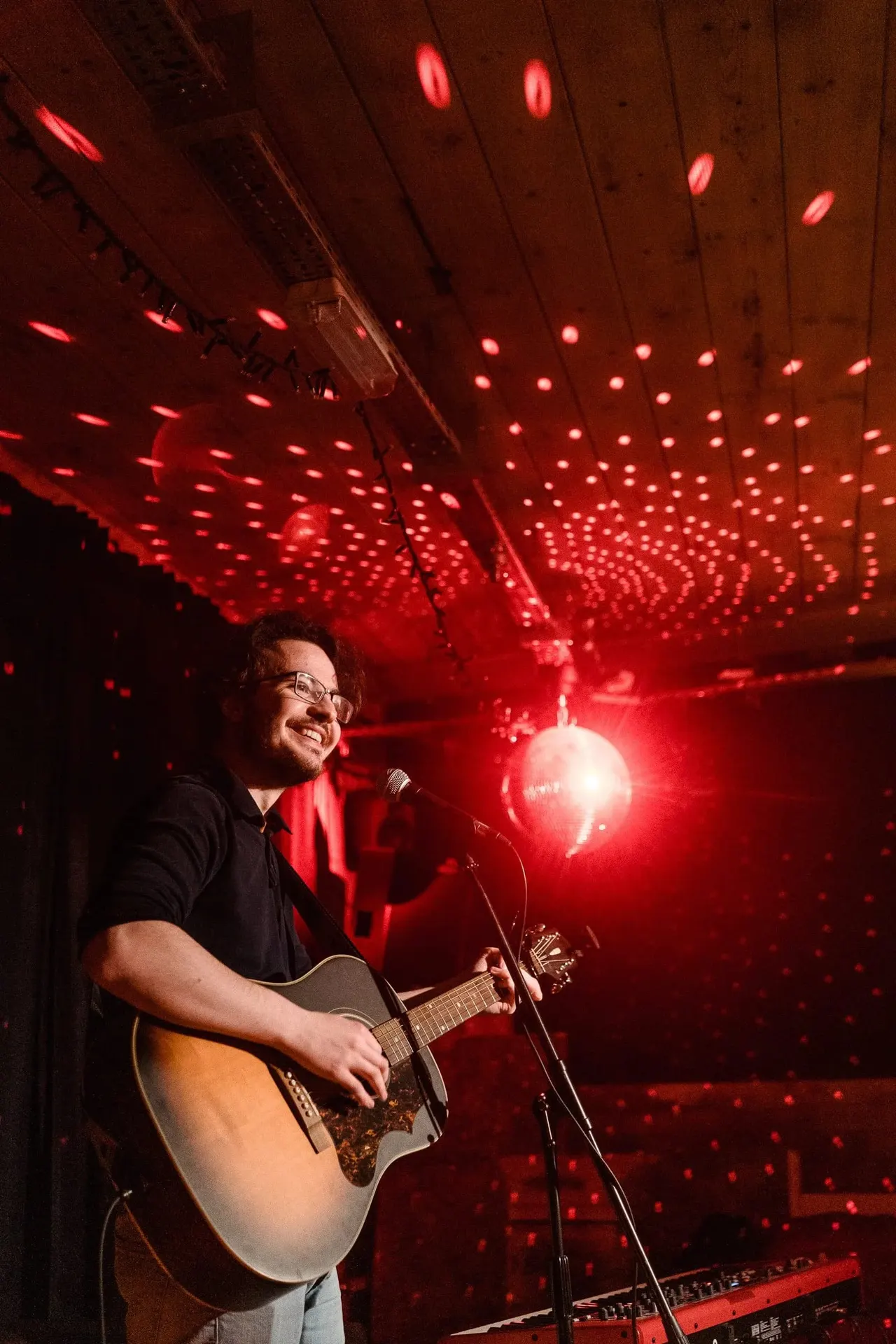 Musician performing with acoustic guitar at a microphone under red stage lighting and illuminated ceiling.
