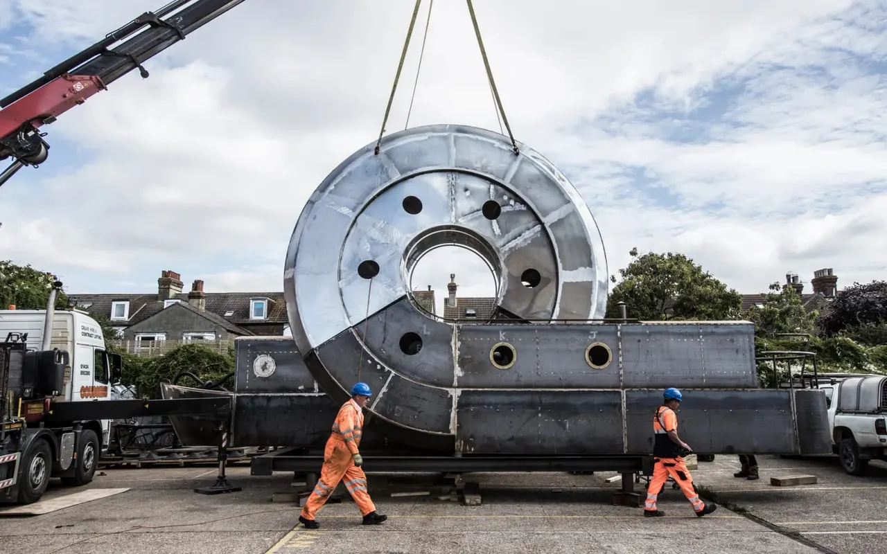 A large metal boiler being hoisted by crane onto a historic steam locomotive, with two workers in orange hi-visibility clothing guiding the operation.