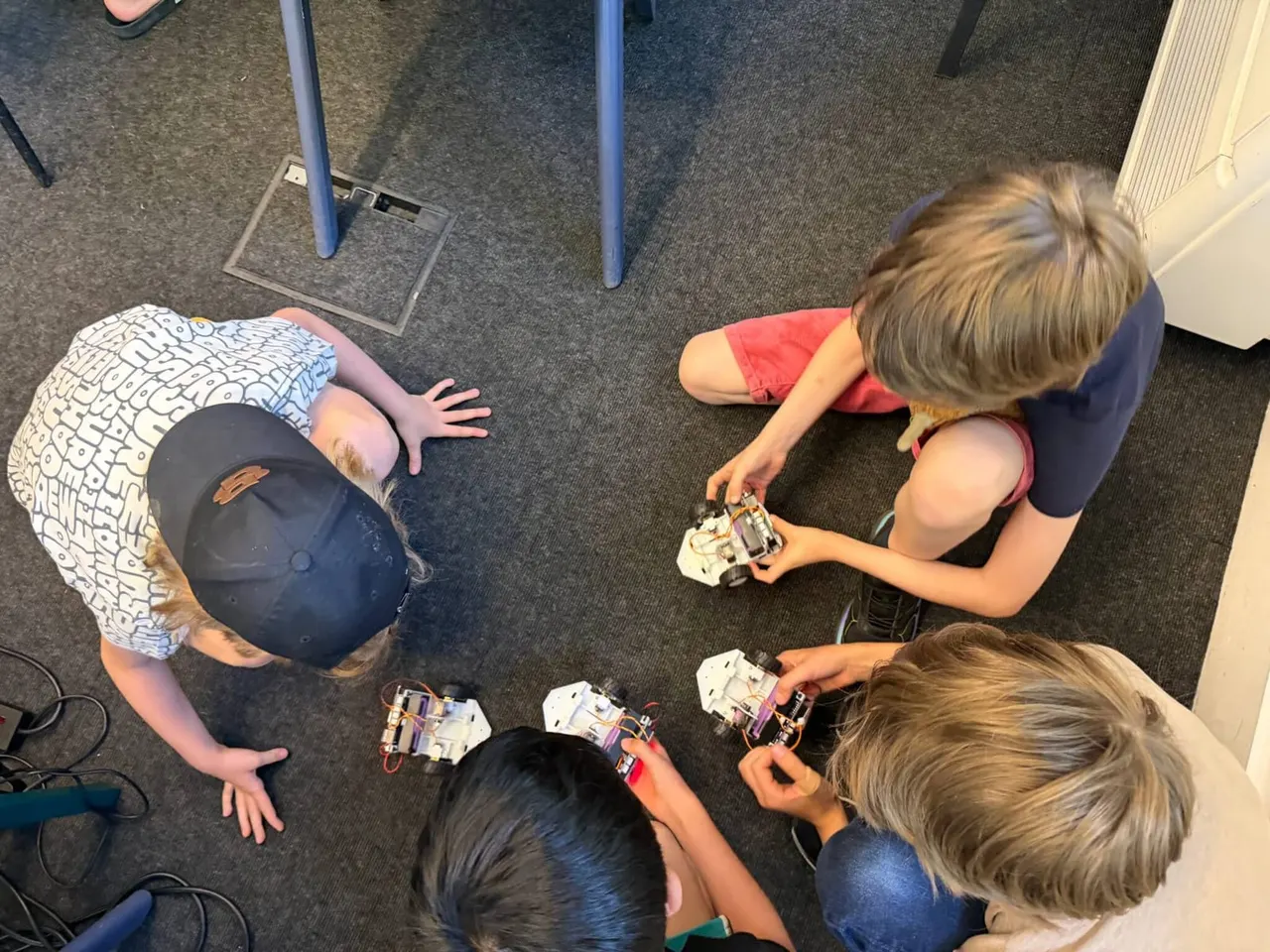 Three young children sitting on grey carpet, examining and handling small electronic circuit boards and components during a robotics workshop.