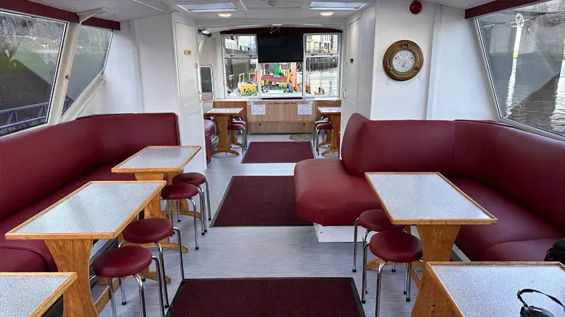Interior of a boat cabin with burgundy seating booths, small tables, and large windows overlooking water.