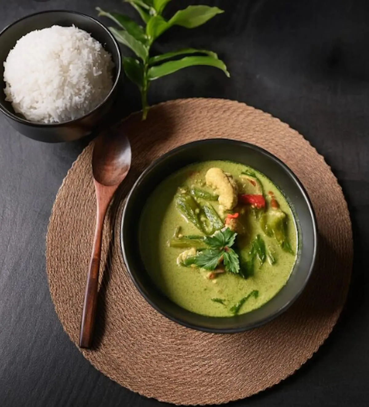Green curry with vegetables and herbs in a bowl, served with white rice and a wooden spoon on a dark surface.