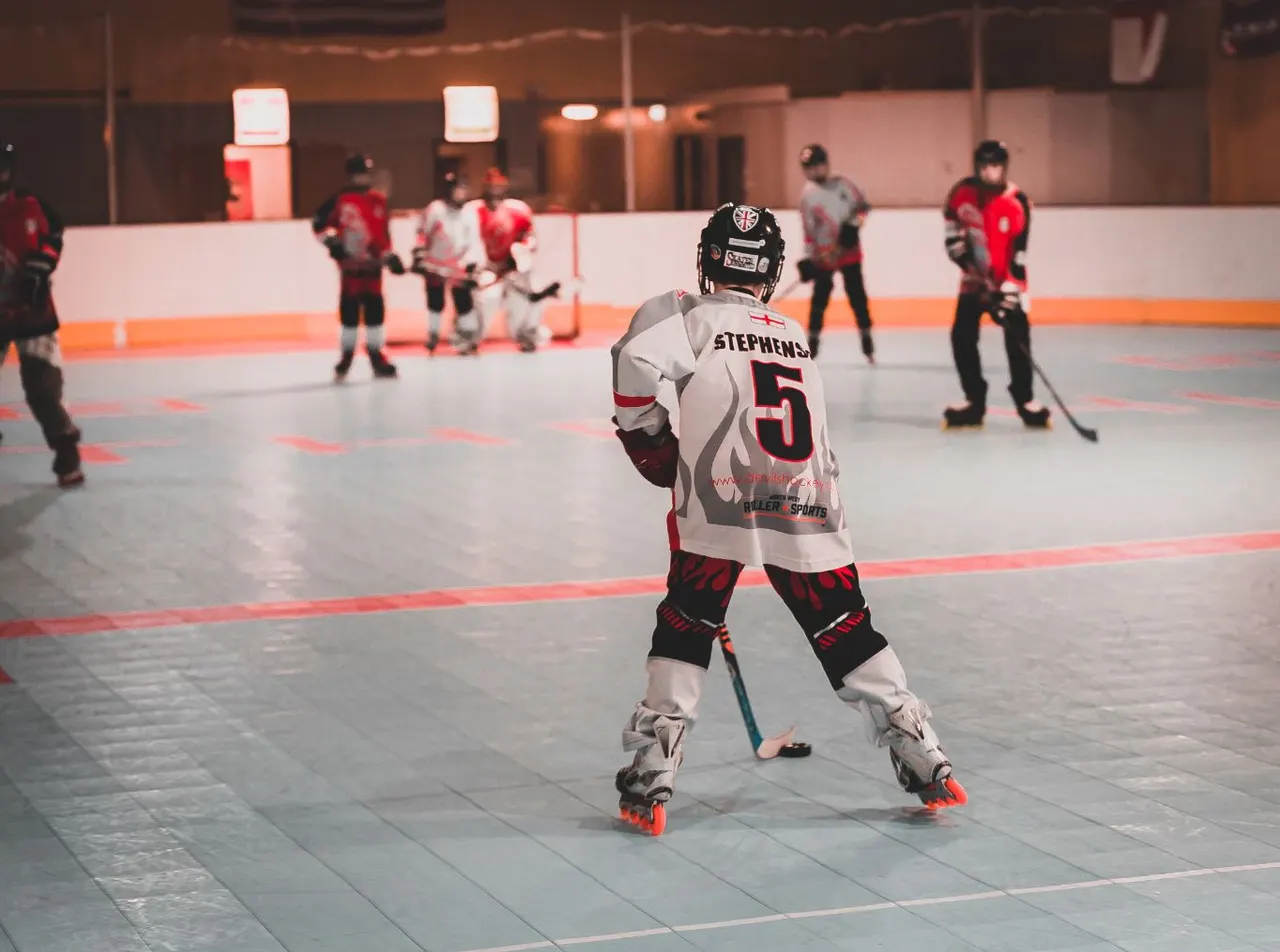 Young inline hockey player wearing number 5 jersey stands with hockey stick on indoor court whilst teammates in red and white uniforms play in background.