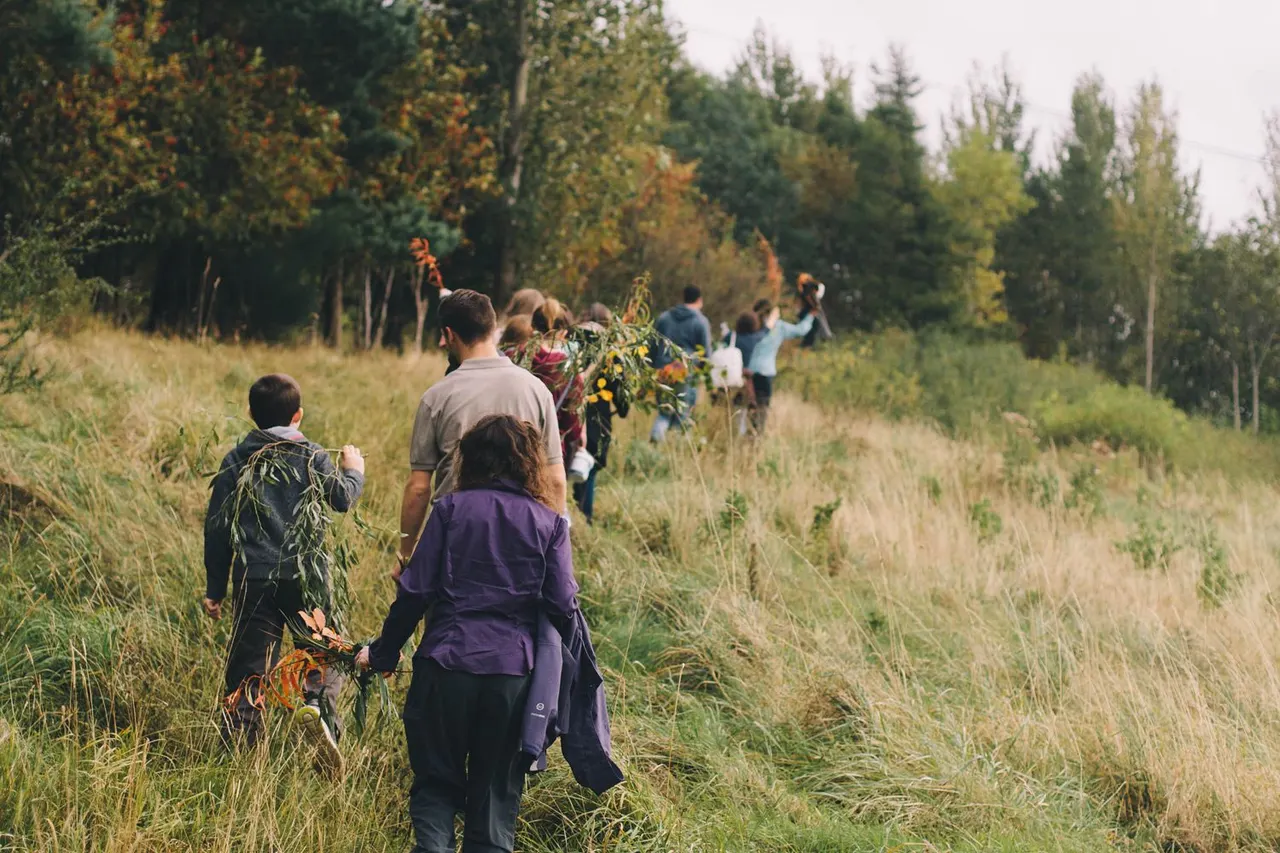 A group of people walking through overgrown grassland with autumn woodland in the background, carrying foraged plants and branches.
