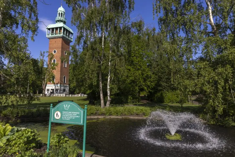 A red brick tower with white lantern top rises behind birch trees, with a fountain spraying in a pond and an information plaque in the foreground.