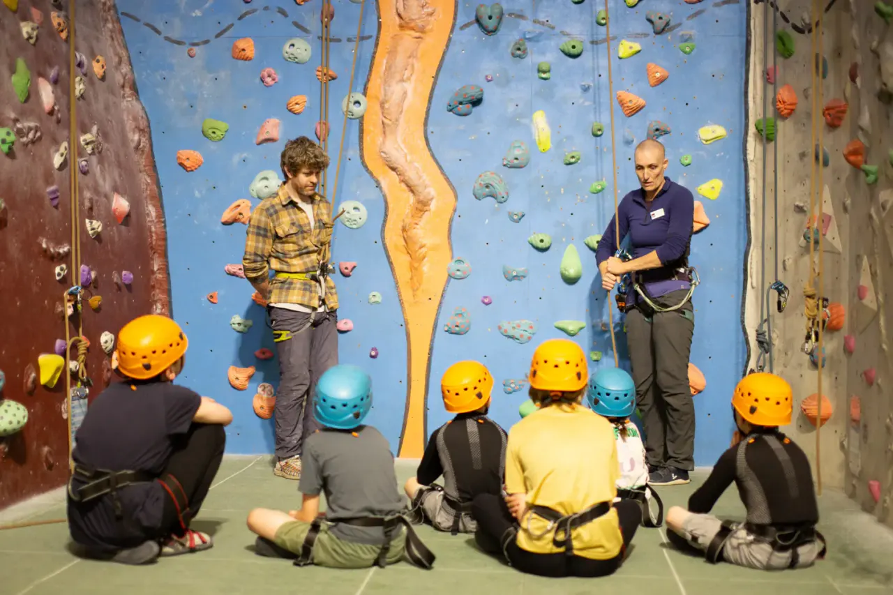 Group of children wearing safety helmets sit facing indoor rock climbing walls whilst two instructors demonstrate climbing techniques.