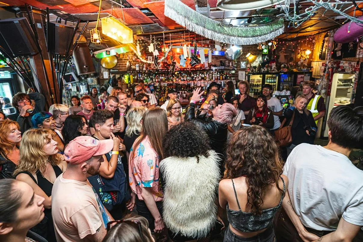 Crowded indoor bar packed with people dancing and socialising, with colourful decorations and a well-stocked bar counter behind them.